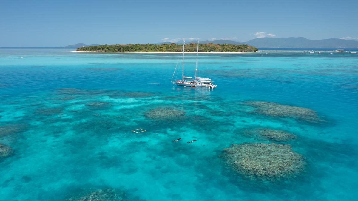 A yacht anchored in clear turquoise water near coral reefs and a tropical island.