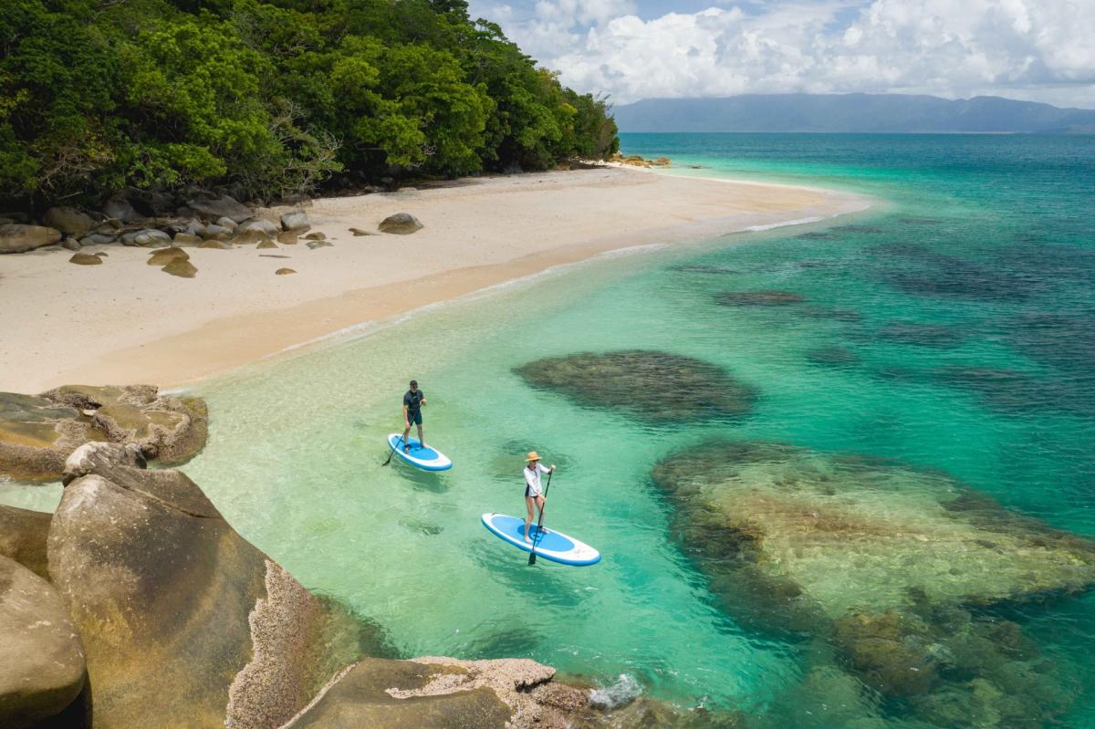 Two people paddleboard in clear turquoise water beside a tropical beach lined with rainforest and granite boulders.
