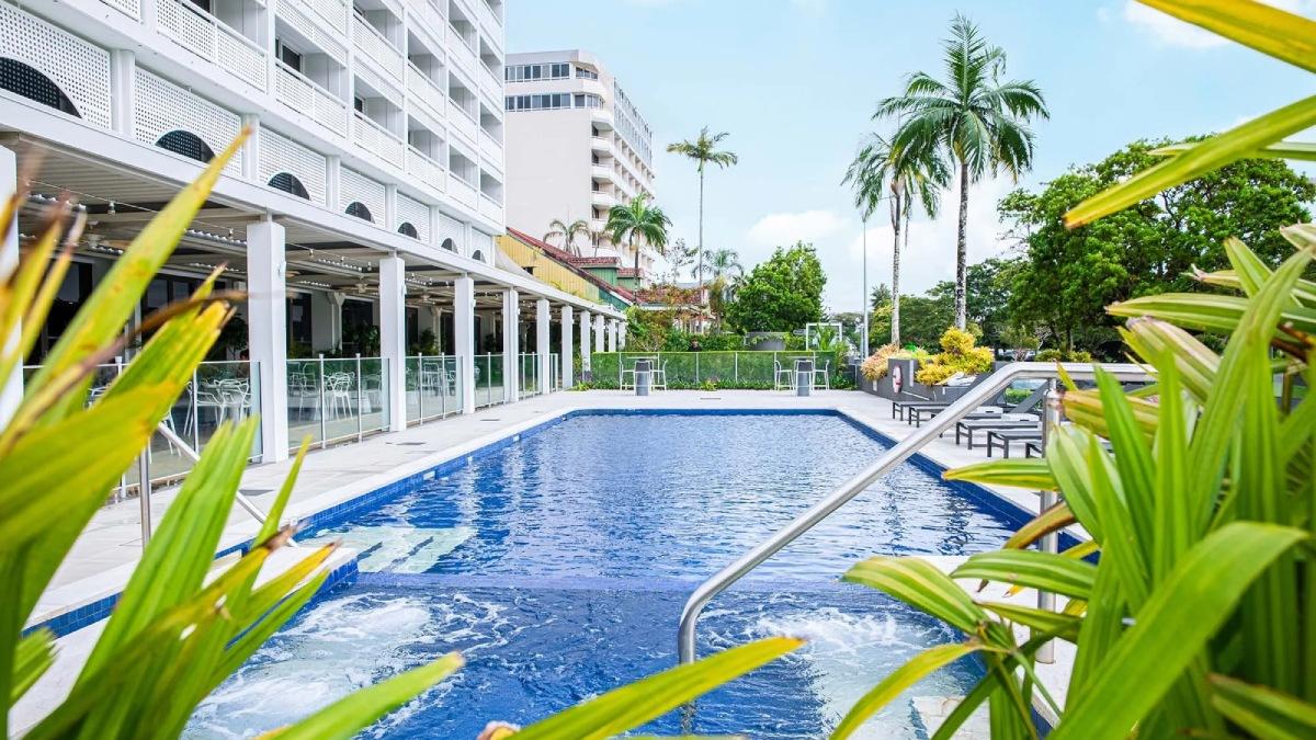 A hotel swimming pool surrounded by palm trees and tropical gardens beside a modern accommodation building.